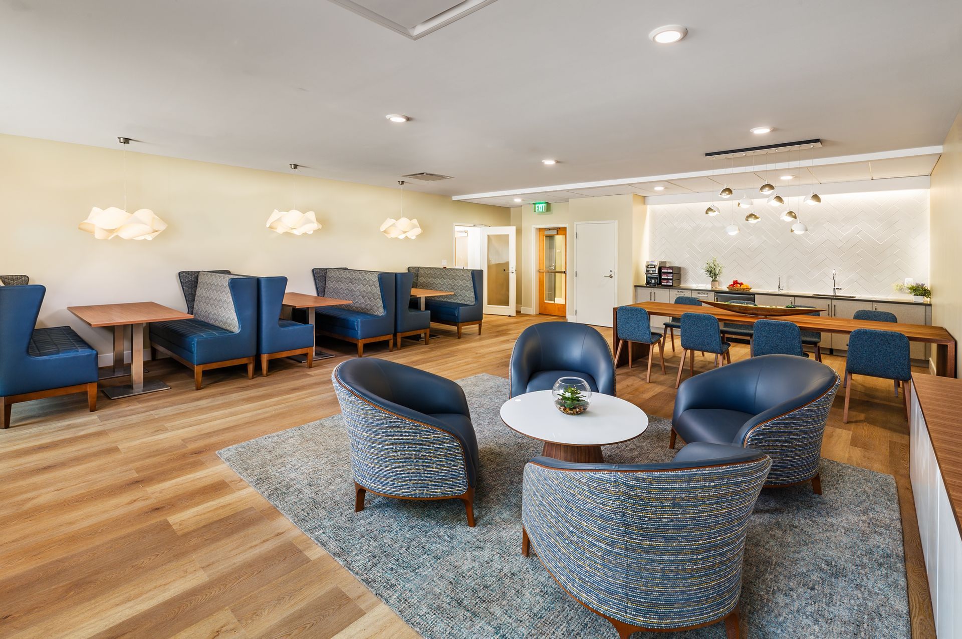 Lounge area with blue seating, wooden tables, and a rug. Features include a coffee station and decorative lighting.