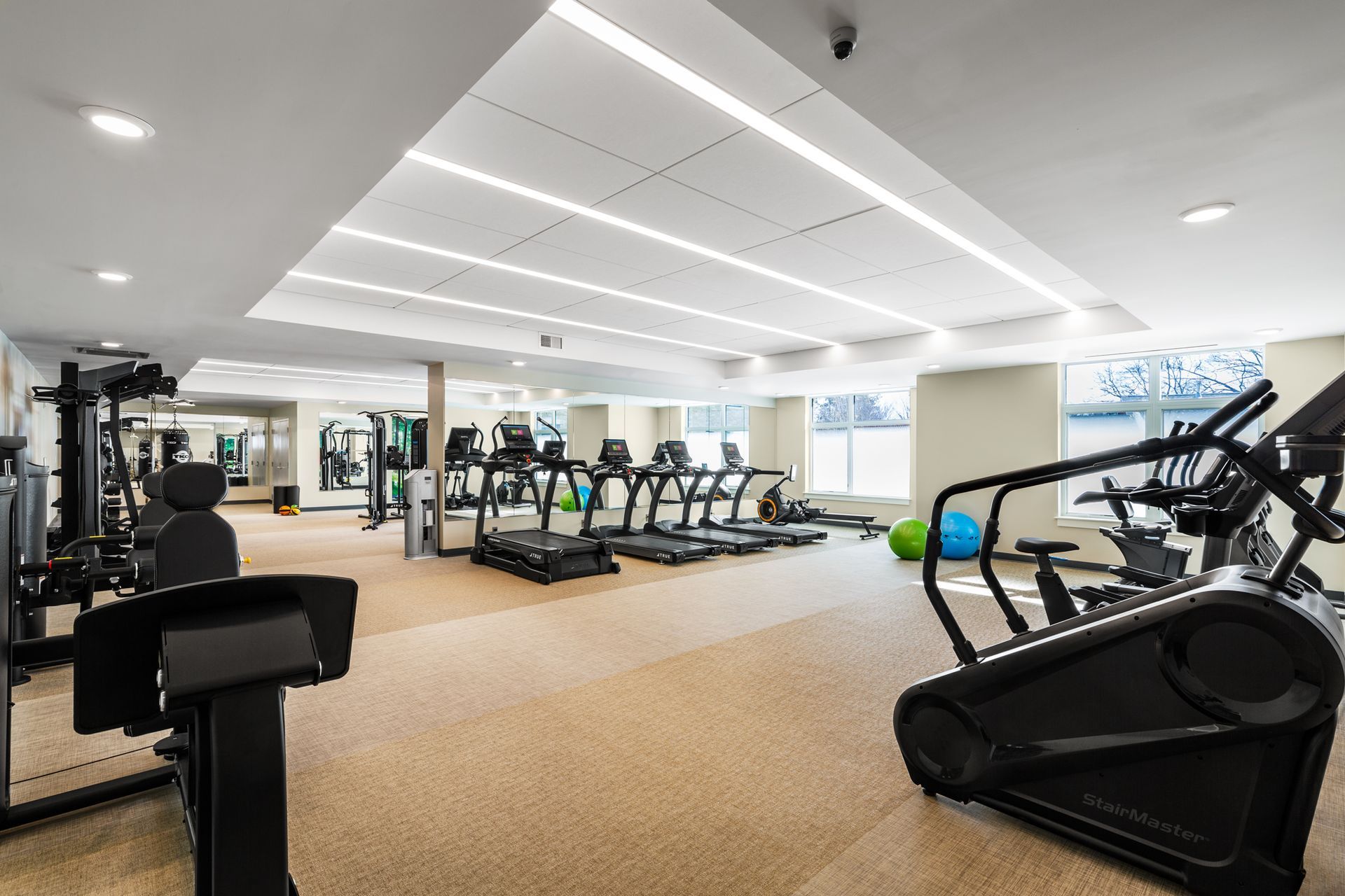 Gym interior with exercise machines: treadmills, weights, and elliptical, tan flooring, bright lighting.