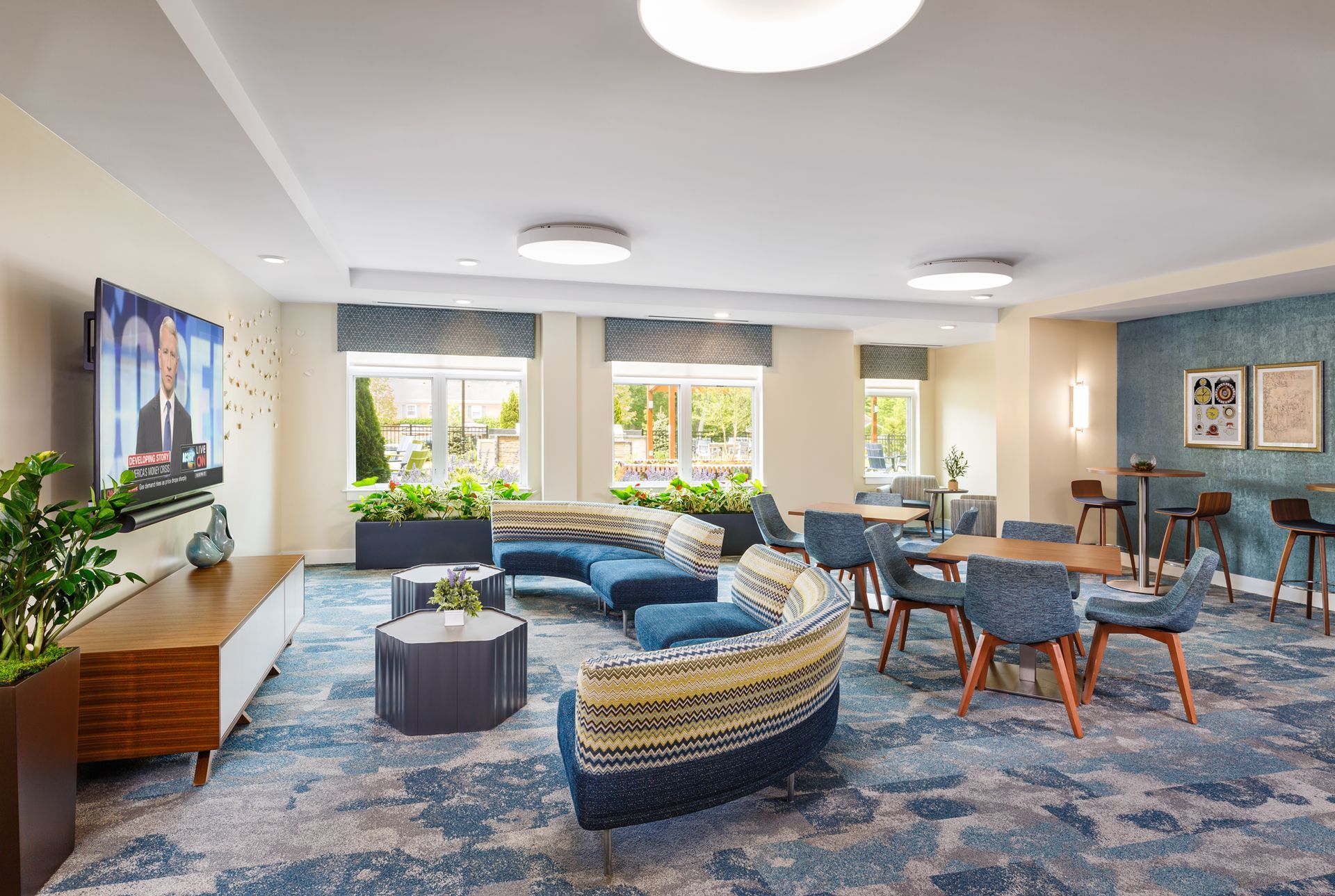 Lounge area with blue patterned carpet, seating, tables, a TV, and windows with outdoor views.