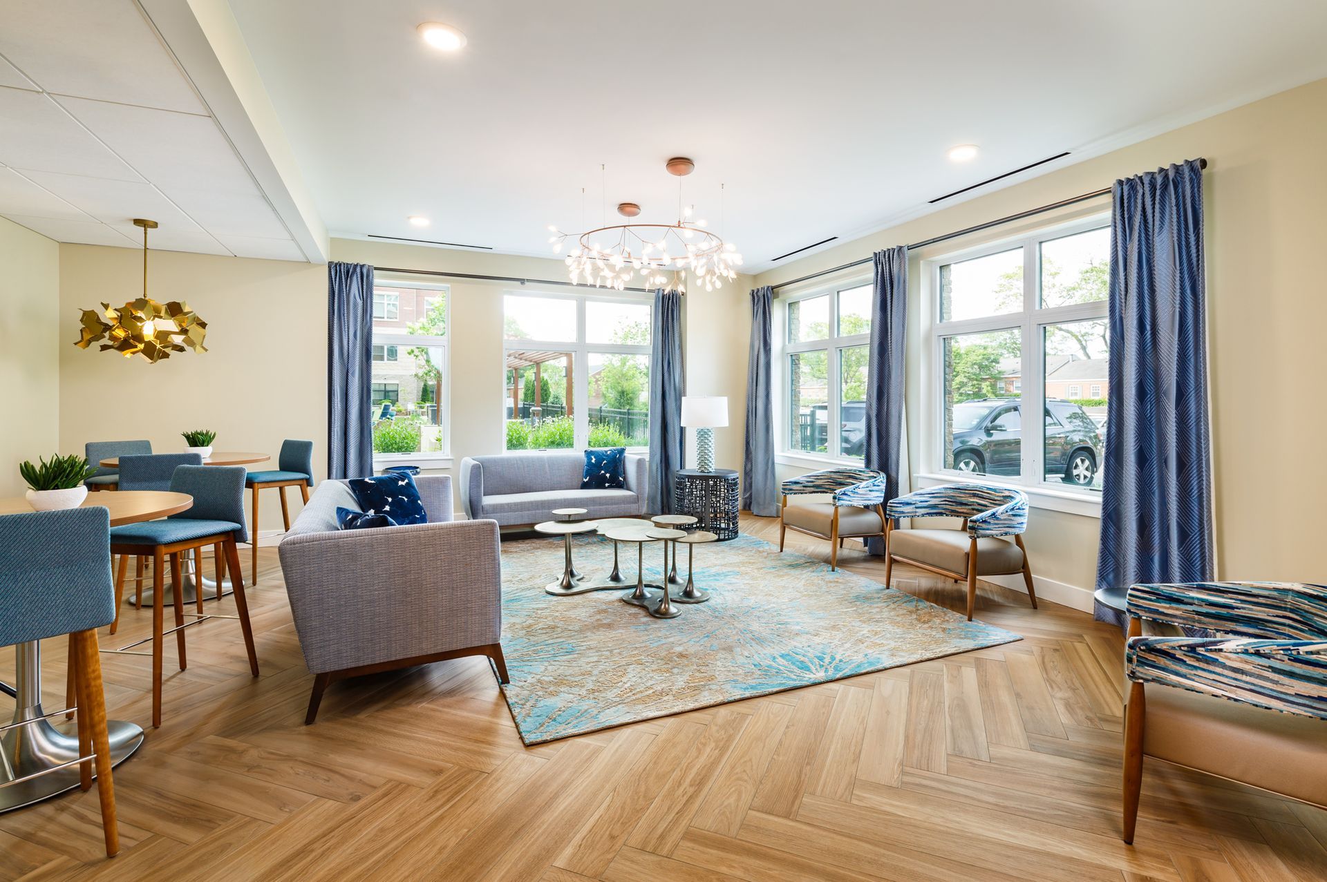 Bright, airy living room with light wooden floors, blue curtains, and a chandelier; includes seating areas.