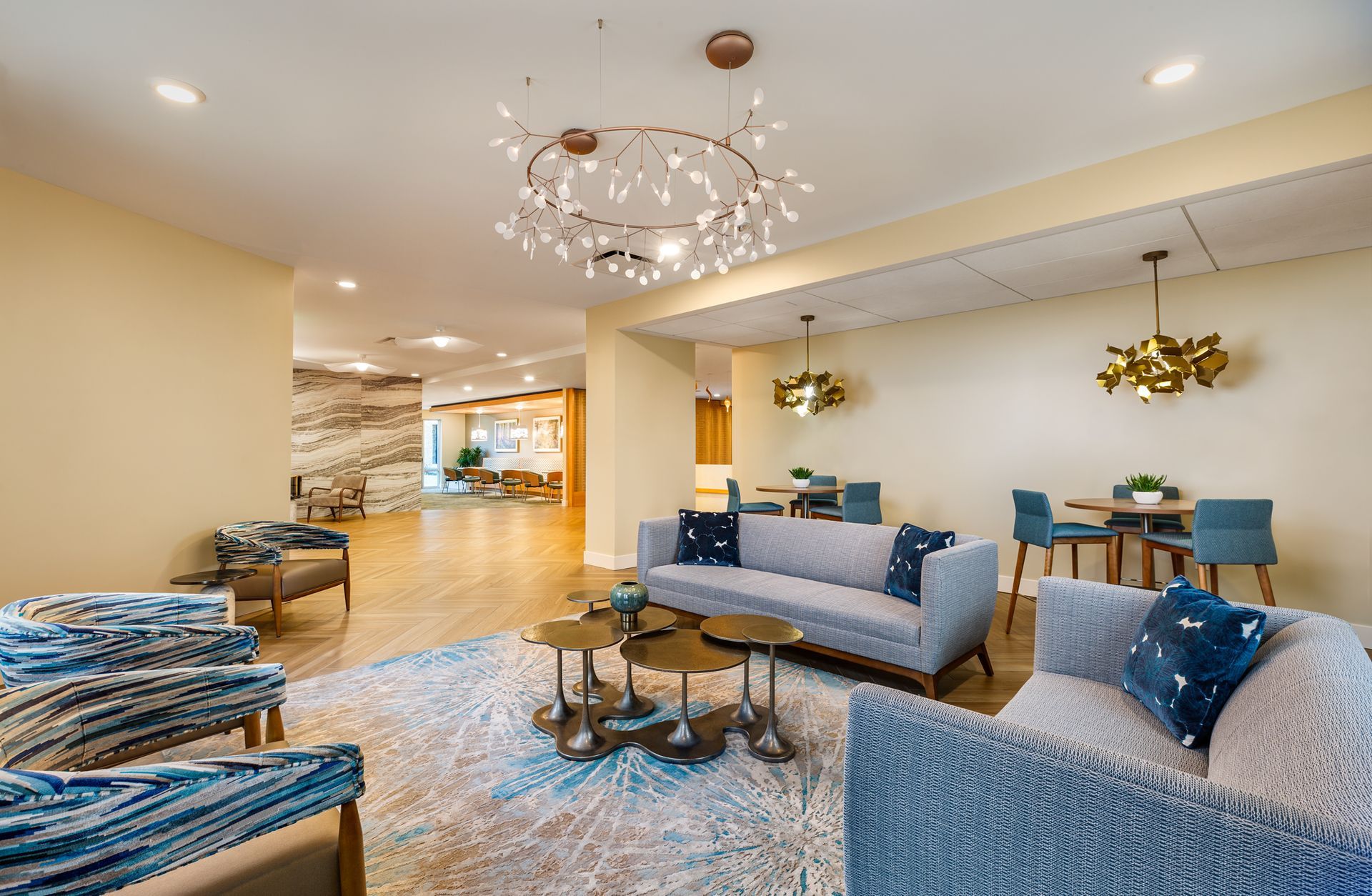 Lounge area with blue sofas, round rug, small tables, and decorative light fixtures. Beige walls and hardwood floors.