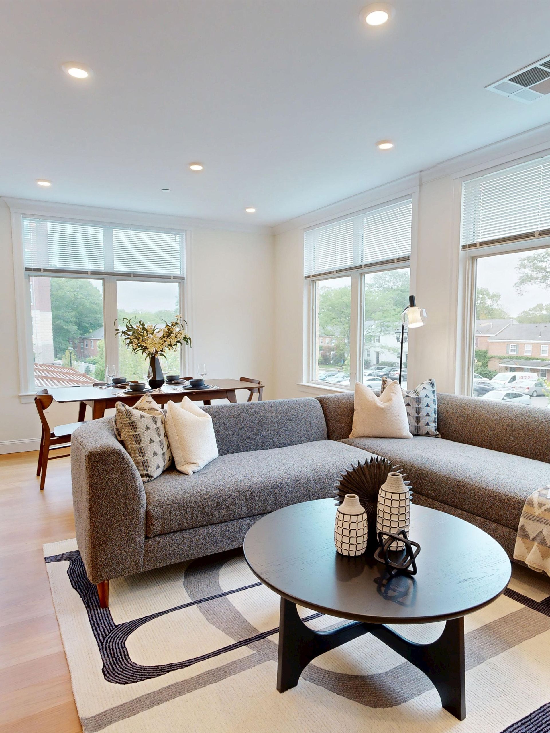 A grey sectional sofa and round coffee table in a sunlit living room, with a dining area visible in the background.