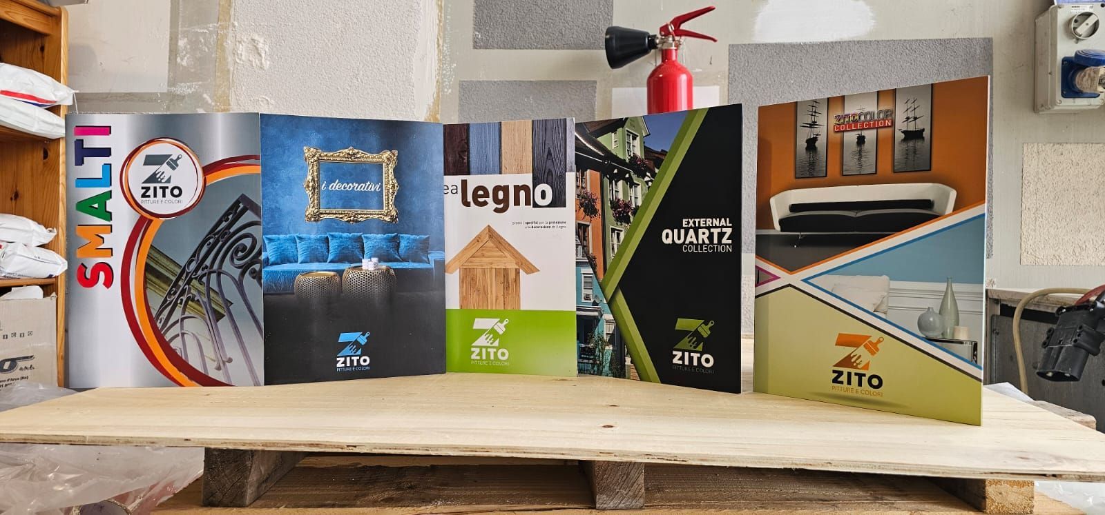 Diversi cataloghi appoggiati su un pallet di legno, con un estintore sullo sfondo.