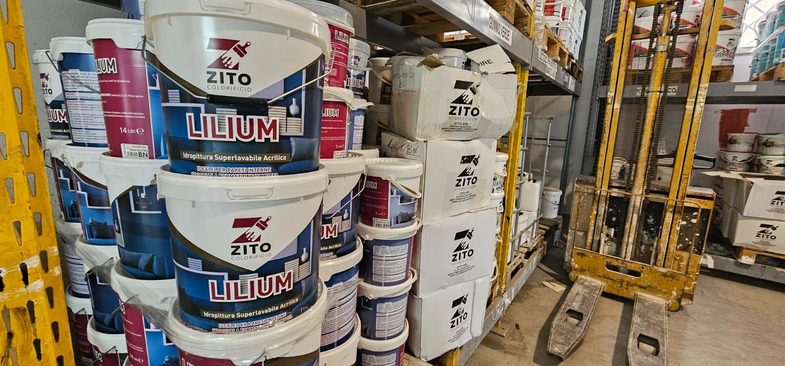 Barattoli di vernice accatastati sugli scaffali di un magazzino; nelle vicinanze c'è un carrello elevatore.