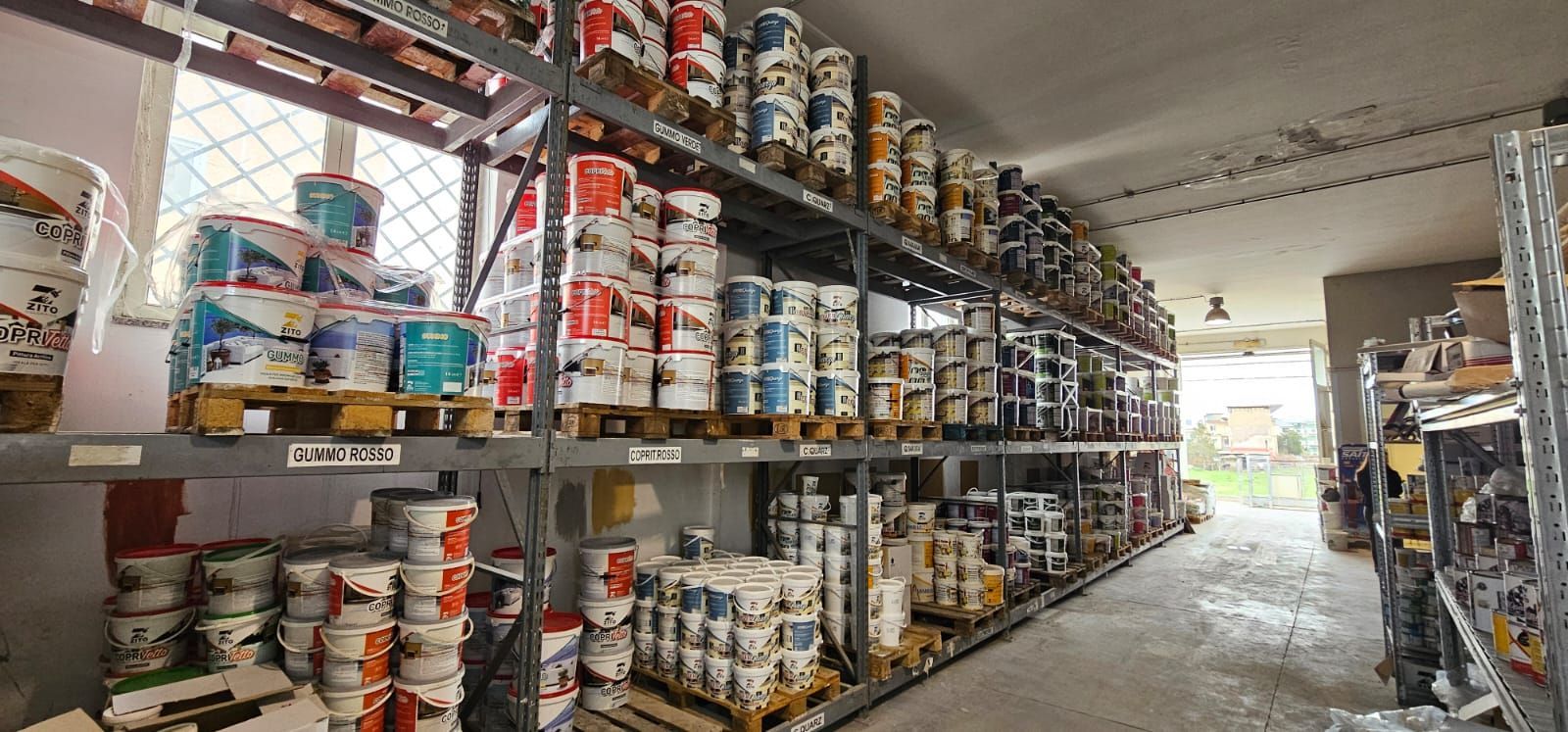 Scaffali pieni di barattoli di vernice in un magazzino, con una finestra e una porta in fondo.