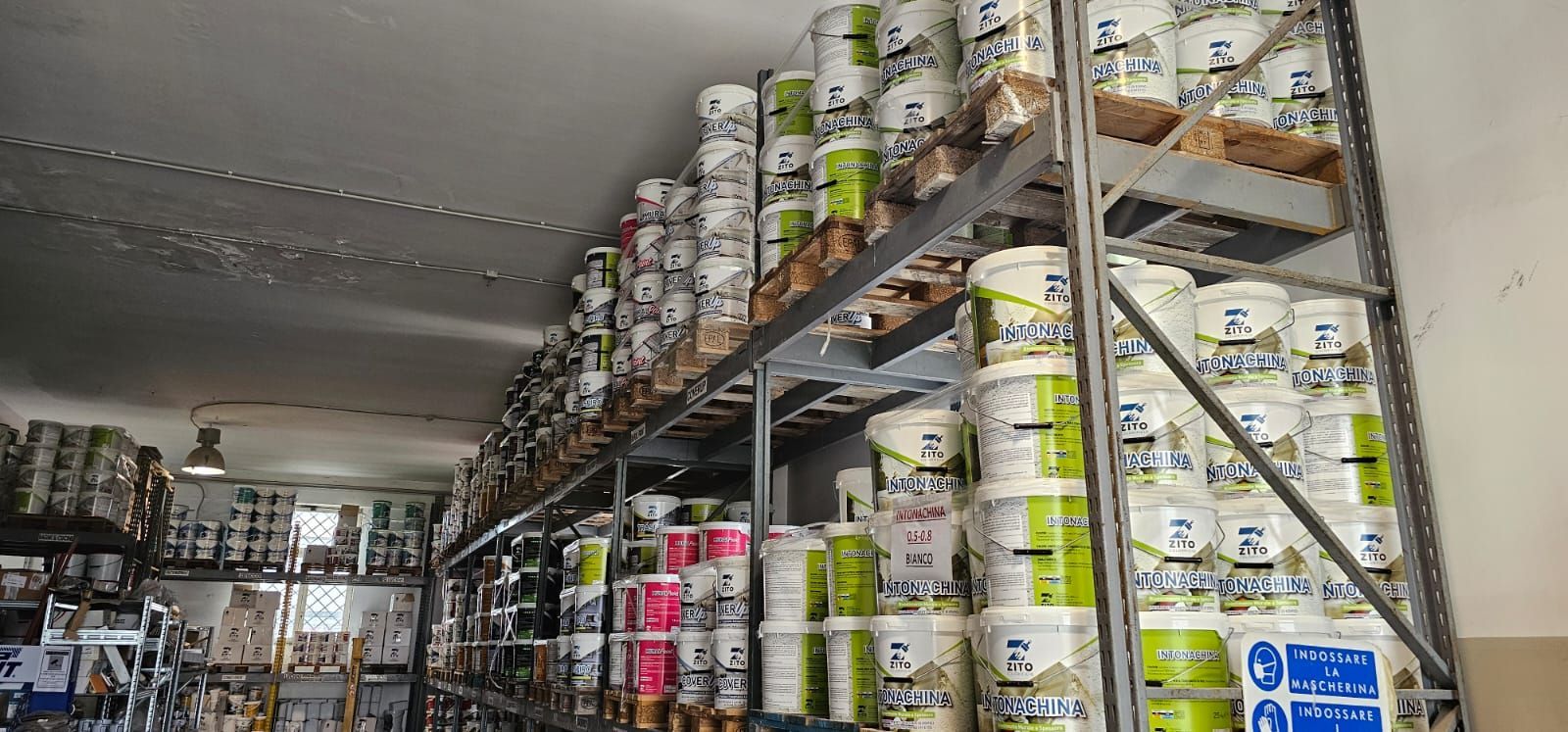 Un'area di stoccaggio piena di secchi di vernice impilati sugli scaffali. I secchi hanno etichette verdi e bianche.