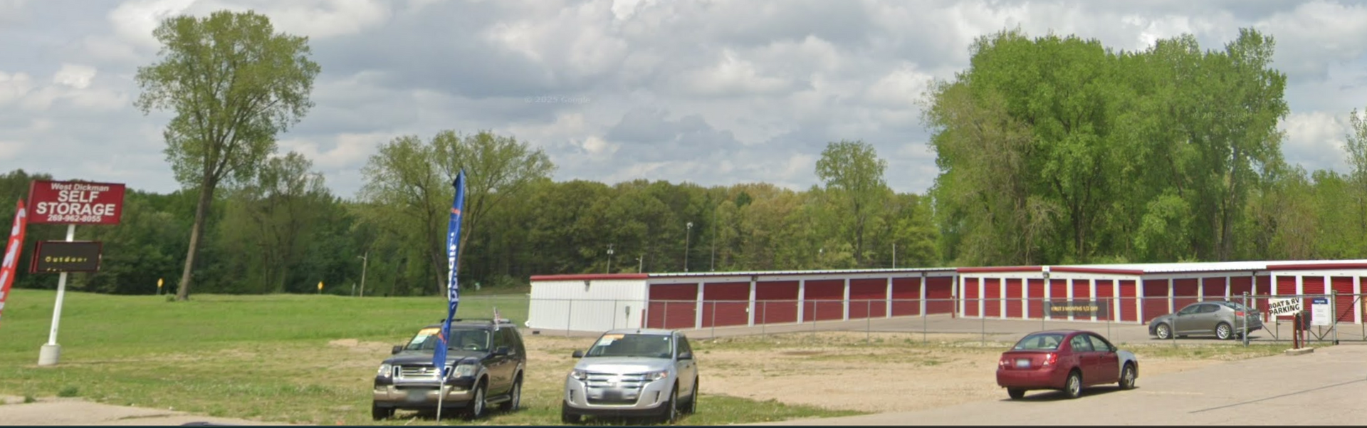 Exterior of storage units with cars parked in front, trees in the background, and a cloudy sky.