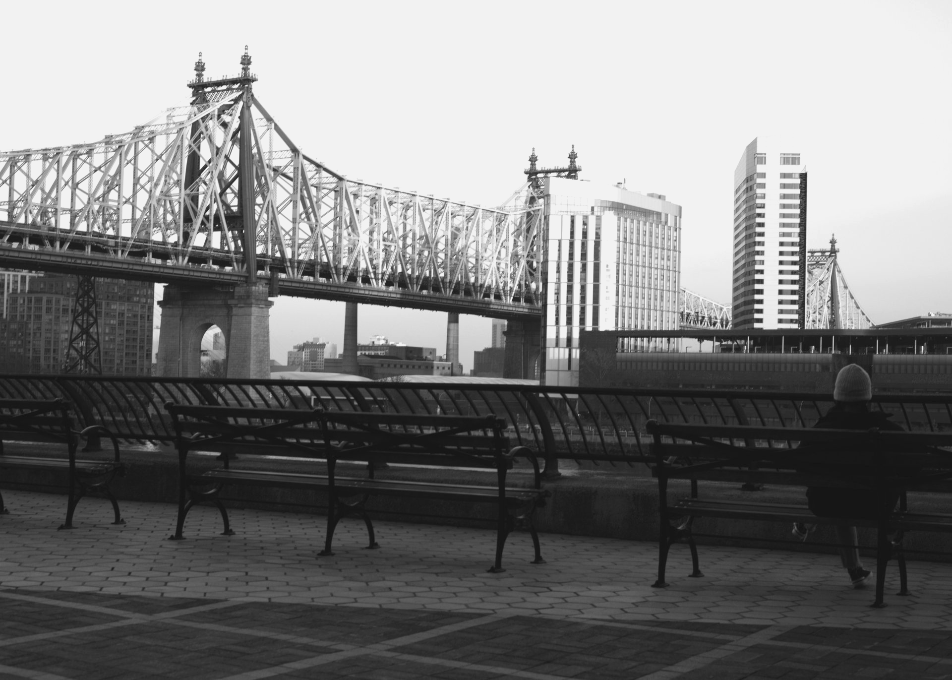 Black and white photo of the 58th Street bridge that connects Manhattan and Queens. On the right of the photo in a park bench sits an unidentified person.