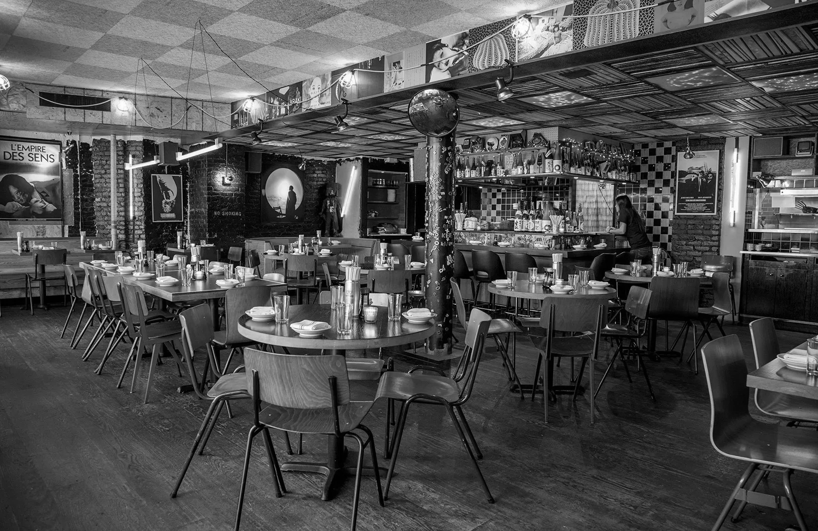a black and white photo of a restaurant dining room, with a bar on the back and tabled on the front.