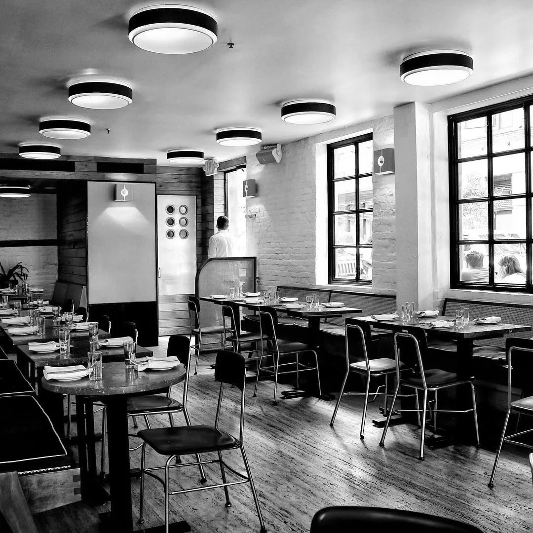 a black and white picture of a restaurant's dinign room, with a male cook looking outside by the door.