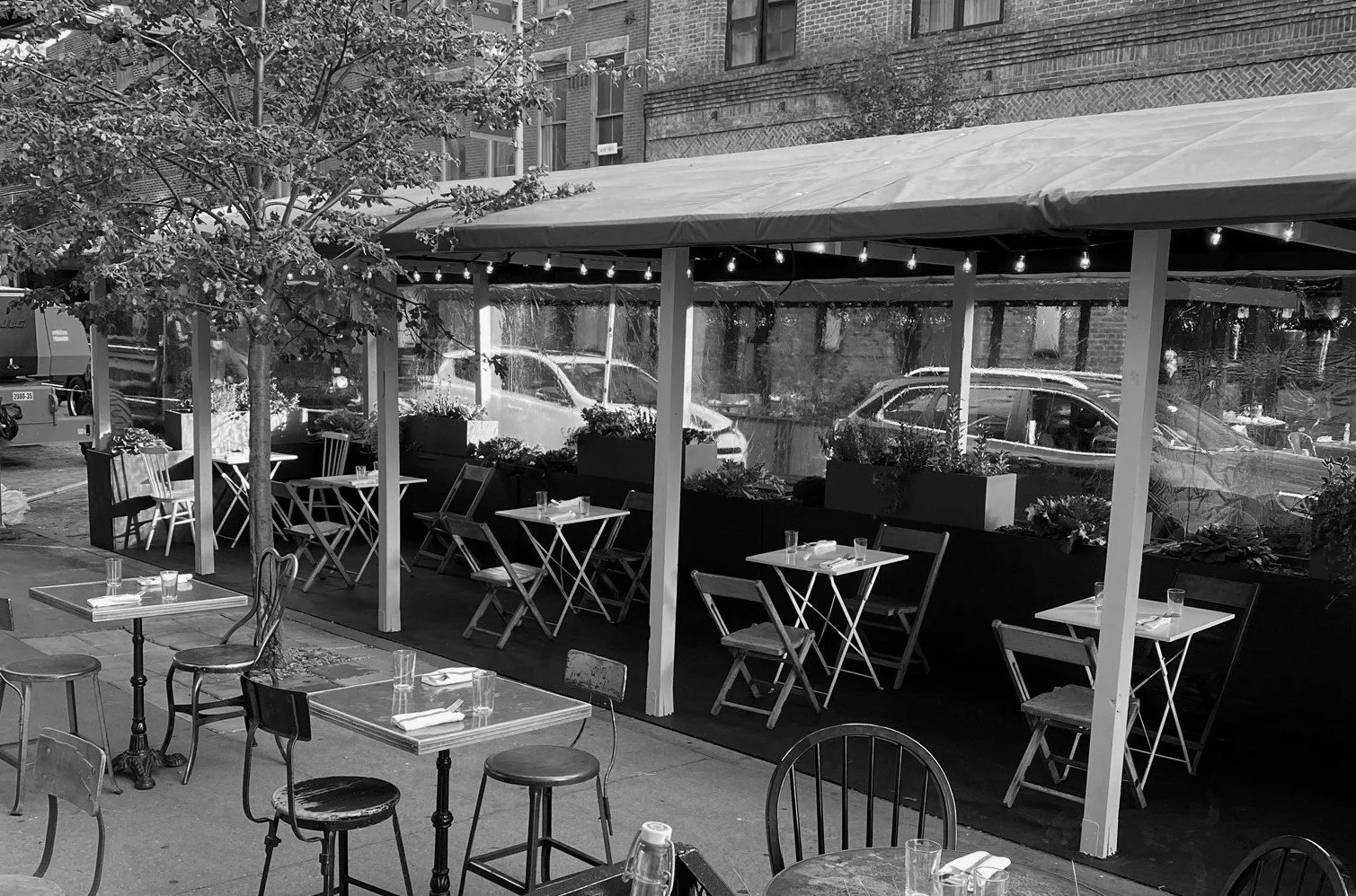 Black and white photo of the outdoor dining area of Yves NYC, featuring 8 tables that seat 2 people each, 5 of which are in a sheed on the street