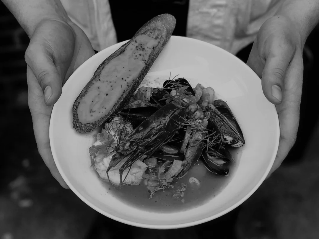 A black and white photo of a seafood plate containing mussels, lobster, prawn, broth, and a piece of toasted bread