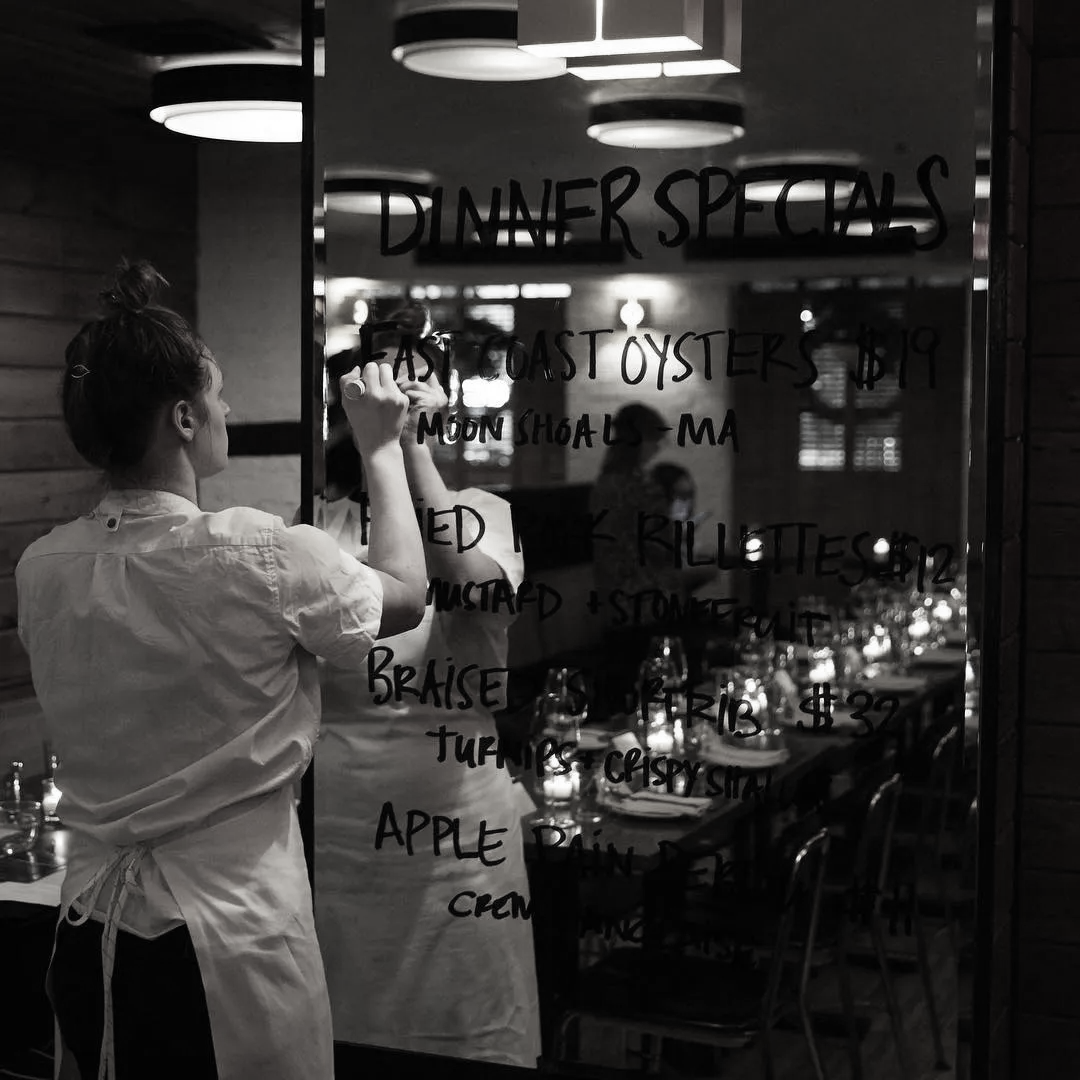 a black and white image of a female server, writing in black marker on a mirror. What she is writing on the mirror is the restaurant's menu.