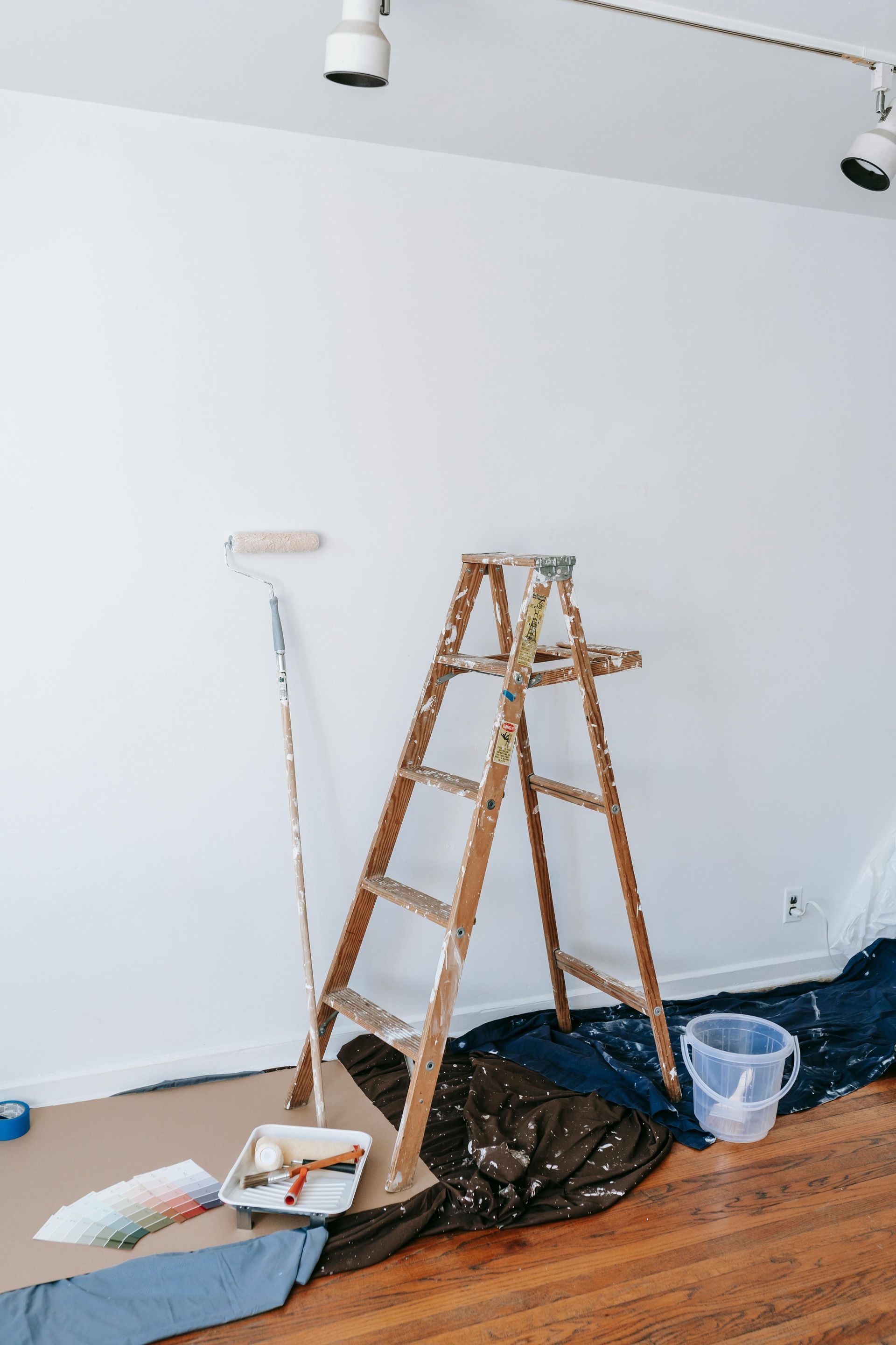 A wooden ladder is sitting on a wooden floor next to a paint roller and bucket.