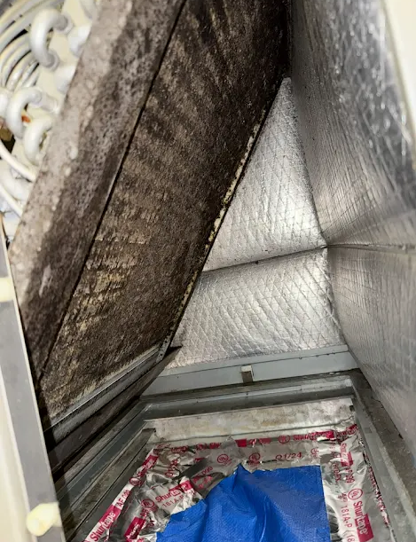Open HVAC duct with silver insulation and a dirty filter panel above a blue item below