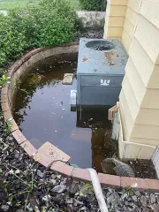 Flooded backyard with standing water around an outdoor HVAC unit near a house wall