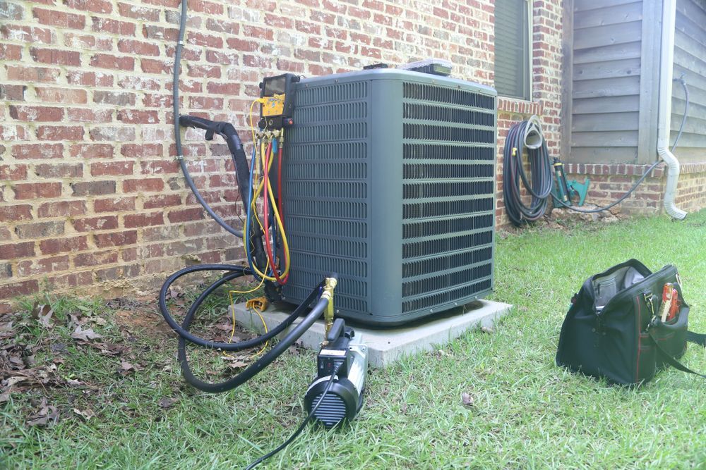 Outdoor air conditioner unit beside brick wall with hoses and tools on grass