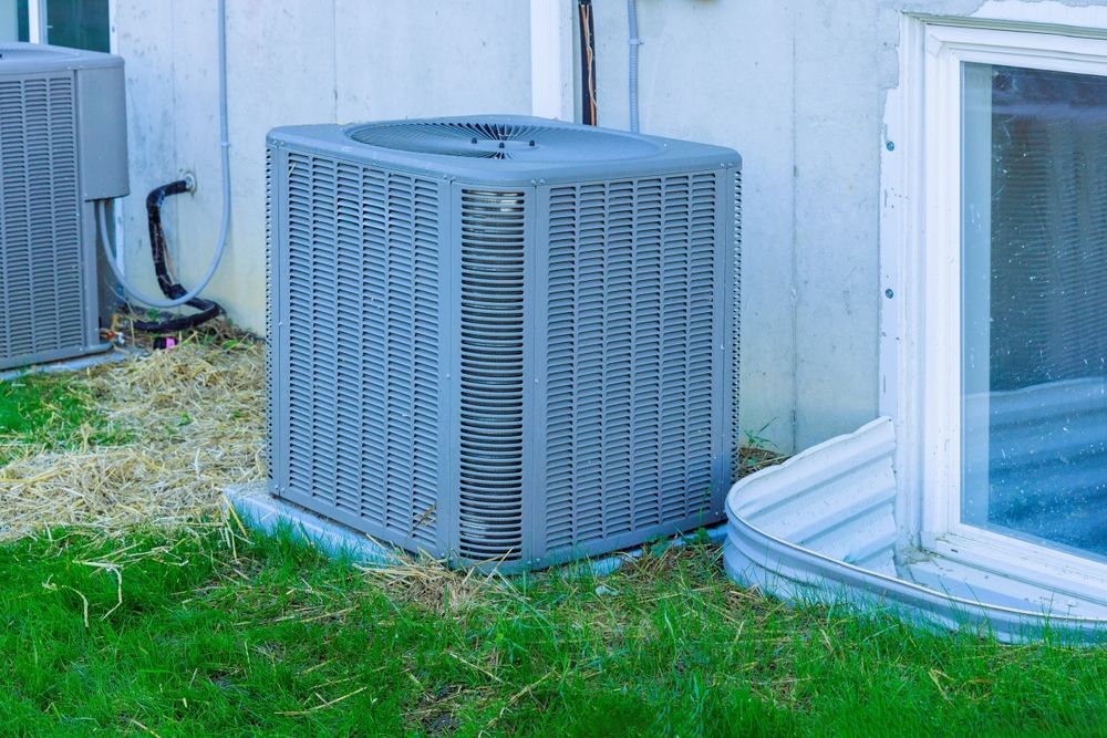 Outdoor HVAC units beside a white house and window, with green grass in front.