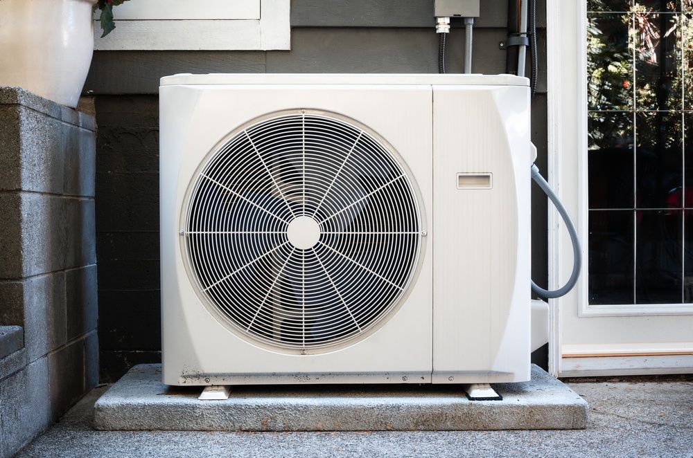 Outdoor heat pump unit beside a building entrance, with a large front fan and white casing