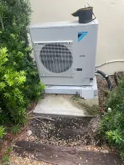 Outdoor air conditioner unit beside a shrub, with pipes and a concrete pad against a wall.