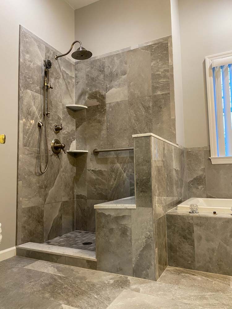 Gray tiled shower with built-in bench, shower head, and handheld shower. Adjacent to a tub.