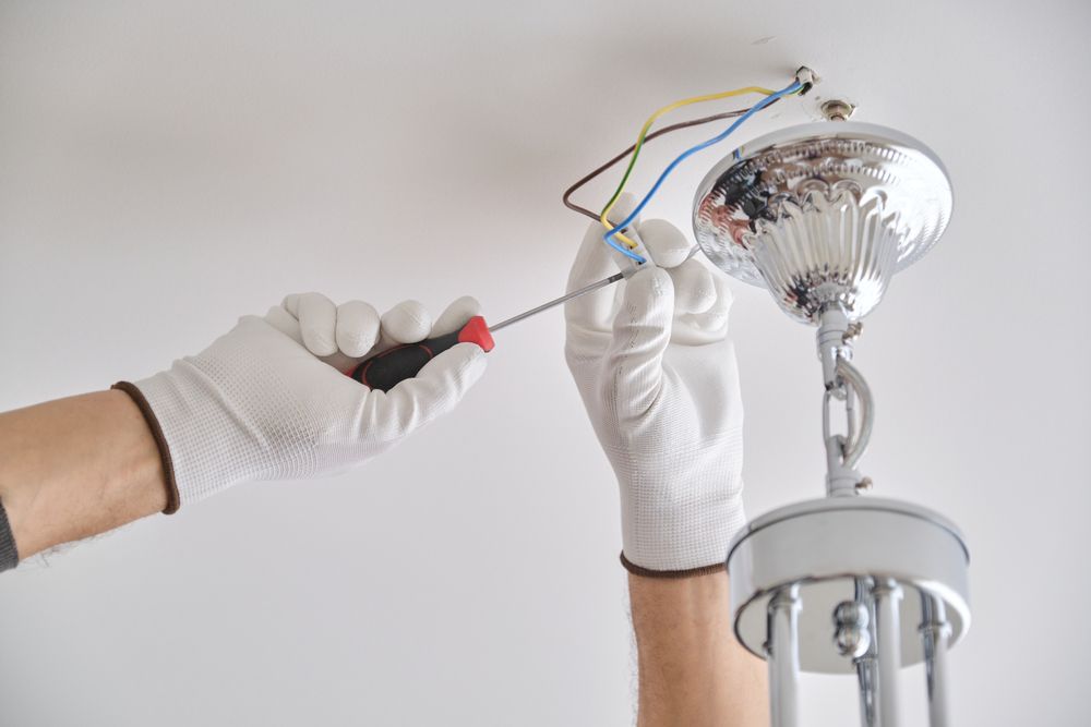 Person wearing gloves installing a chandelier, using a screwdriver, wires exposed.
