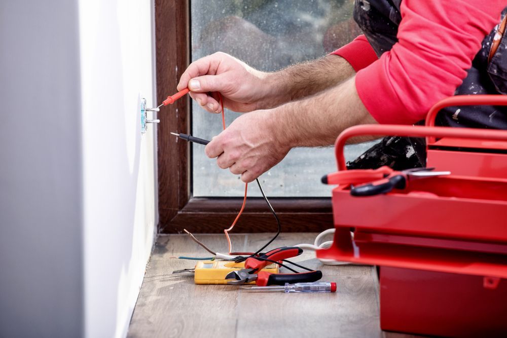 Electrician using a multimeter on a wall outlet, red toolbox nearby.