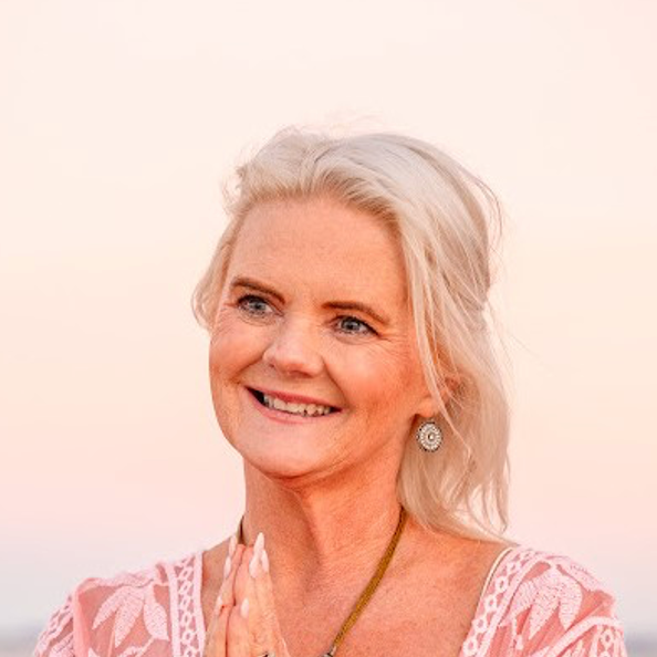 A woman with blonde hair is smiling and praying on the beach.