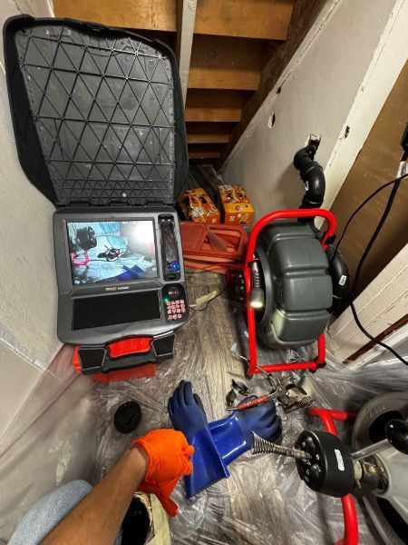 Plumber in crawl space using camera and drain cleaning equipment. Dark screen, blue and orange gloves.