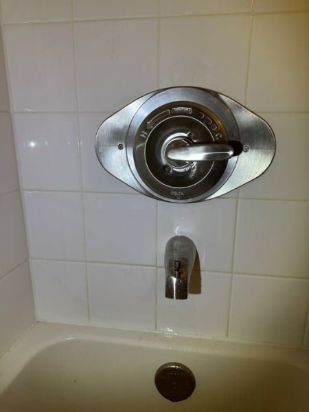 Shower faucet with a silver handle, surrounded by a metal plate, on white tiled wall above a bathtub.