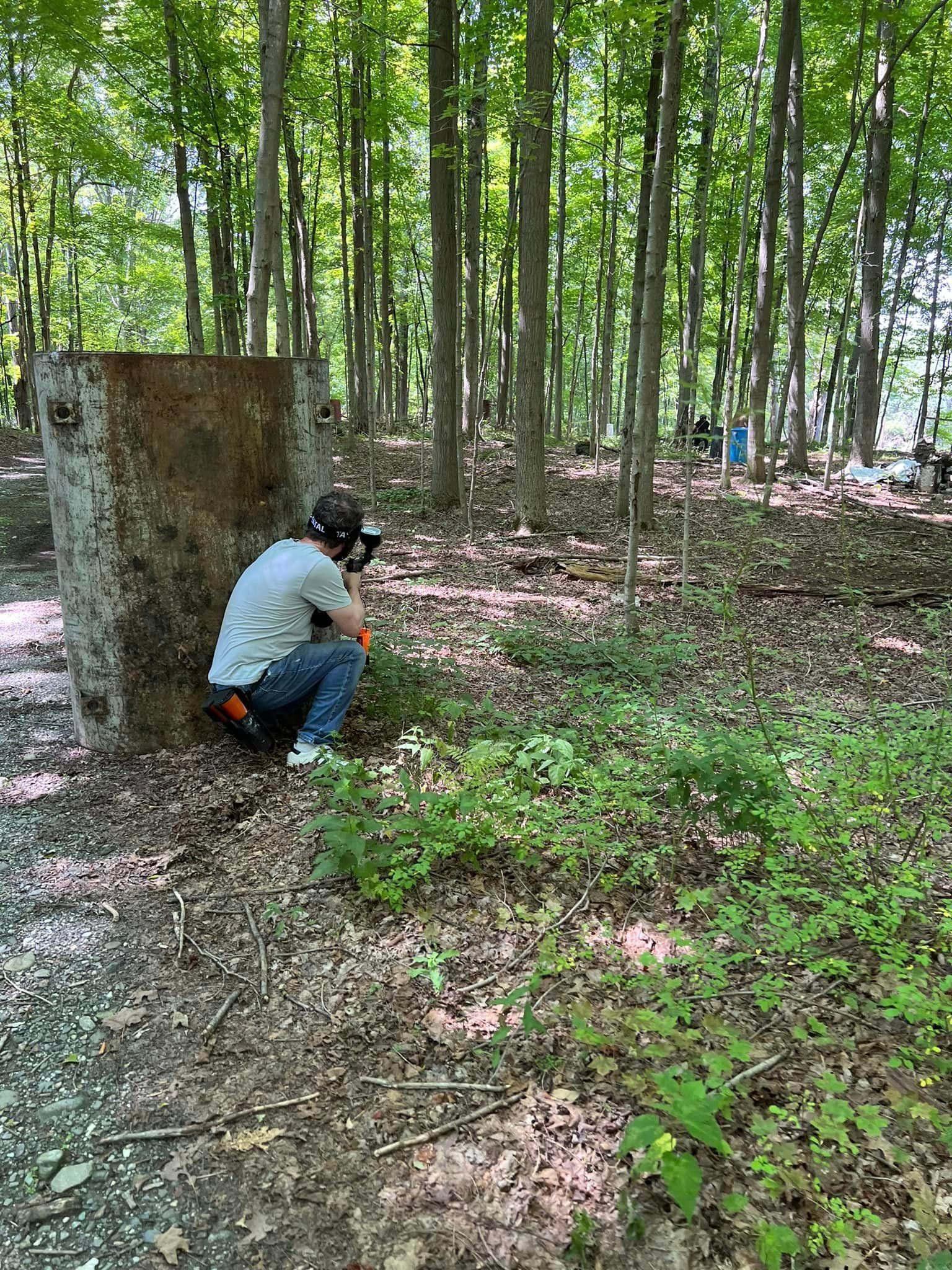 Person in paintball gear crouches behind a concrete barrier in a wooded area, aiming a paintball gun.