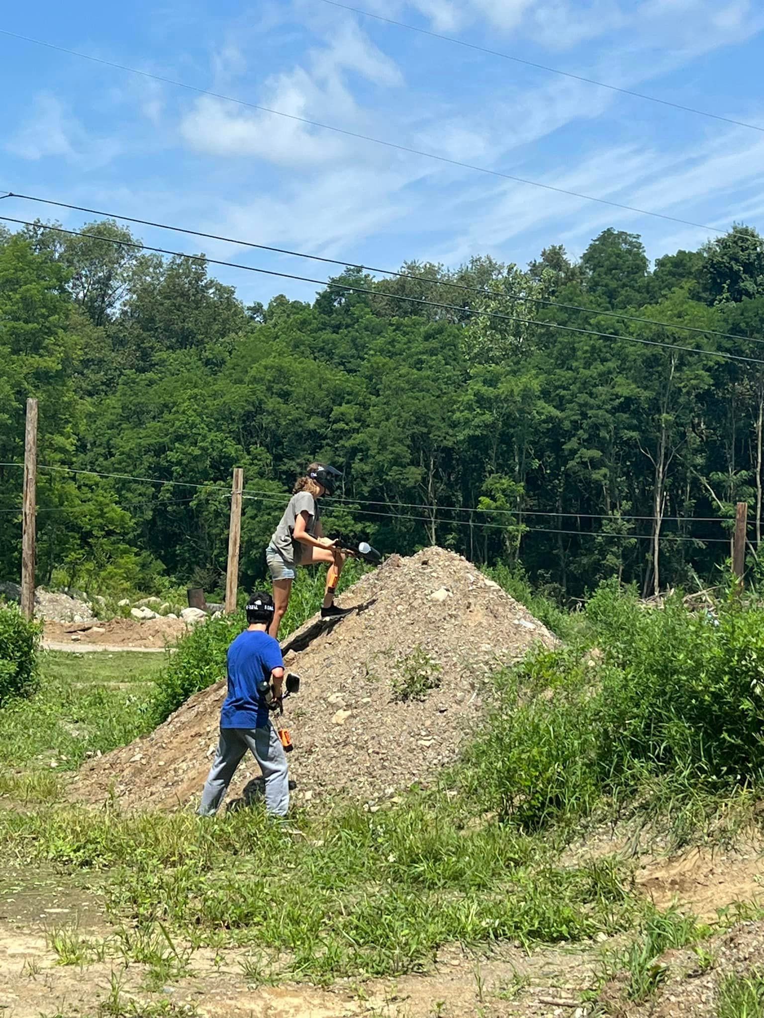 Two people on a dirt mound; one films as the other stands with a ball near trees under a blue sky.