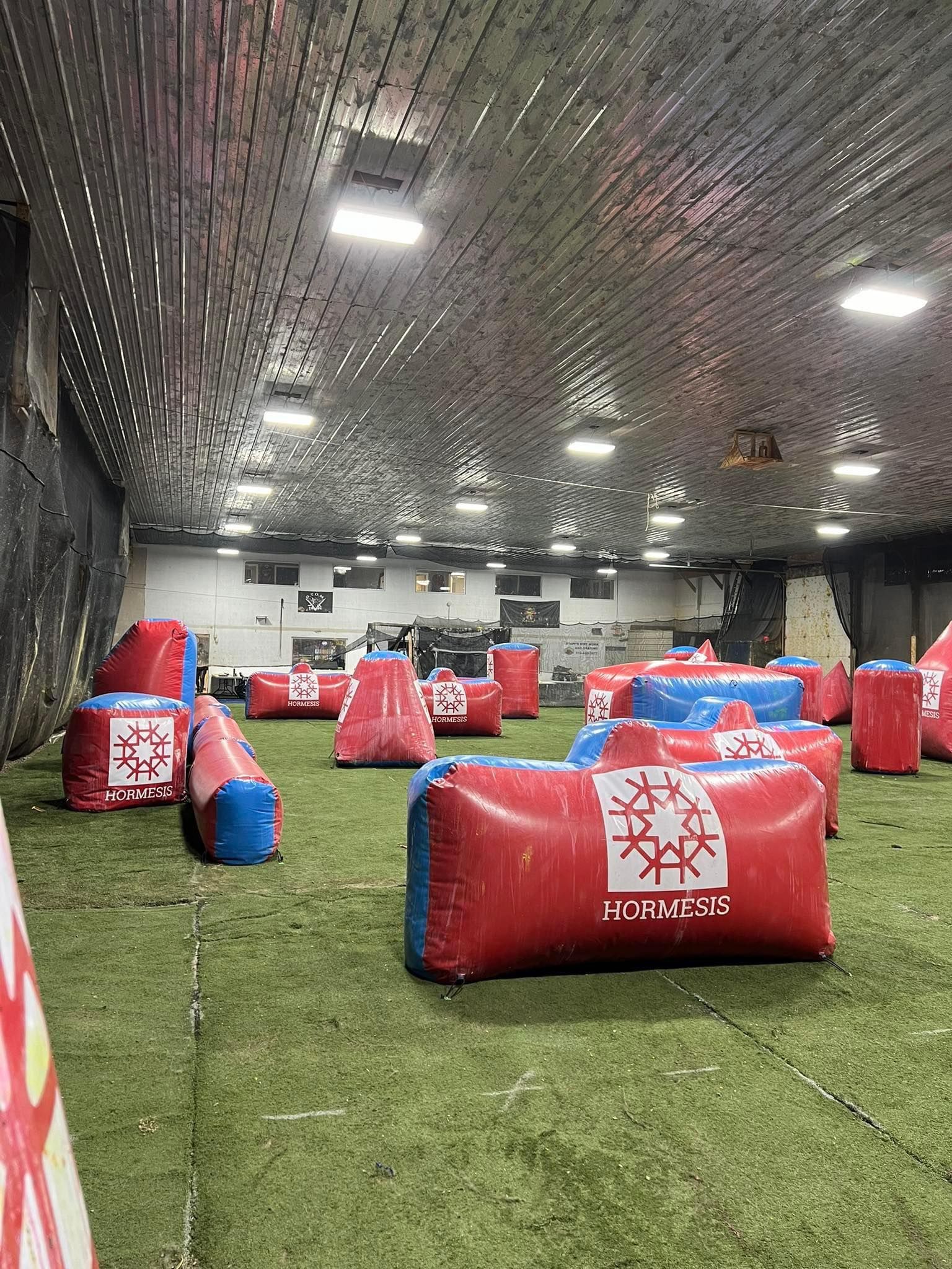 Indoor paintball field with red and blue inflatable bunkers on artificial turf.