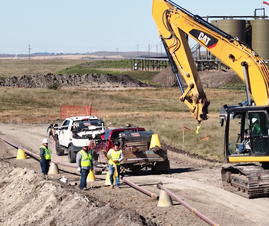Workers in high-visibility vests supervise a pipeline construction site near a yellow CAT excavator and utility trucks.