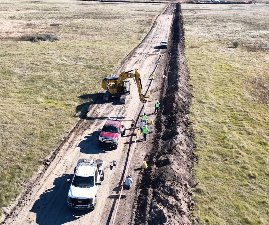 An aerial view shows workers and a yellow excavator installing a pipeline along a trench in a grassy, open field.