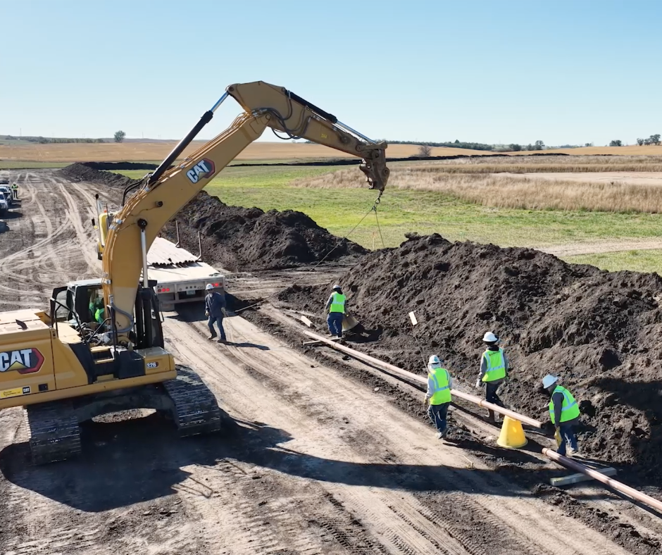 A yellow CAT excavator suspends a pipe over a dirt construction site as workers in high-visibility vests look on.