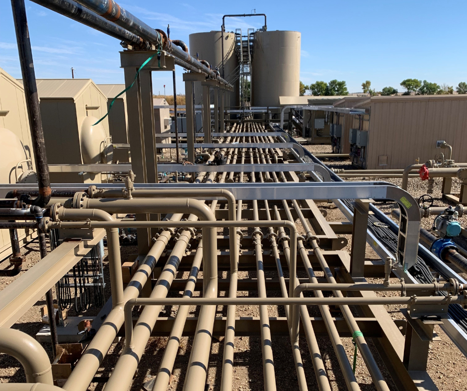 An industrial oil and gas facility with multiple beige tanks, pipes, and metal conduits under a clear blue sky.