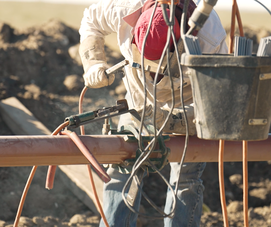 A welder in protective gear works on a pipeline in a dirt field.