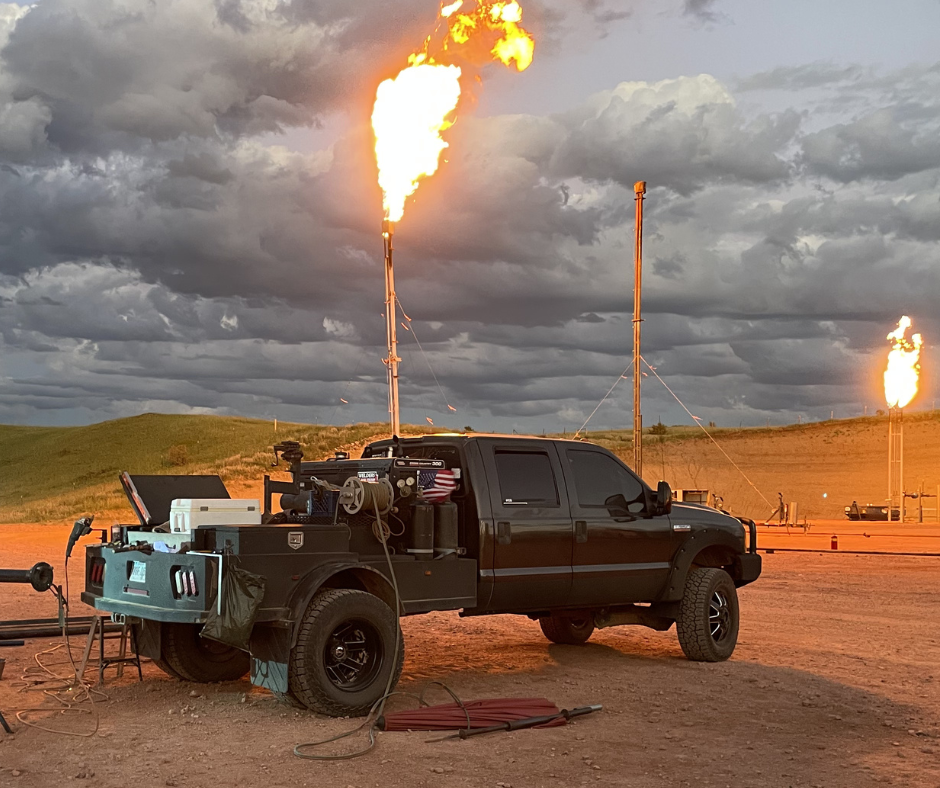 A black pickup truck parked in an oil field at dusk, with a tall flare stack burning bright orange flames above the bed.