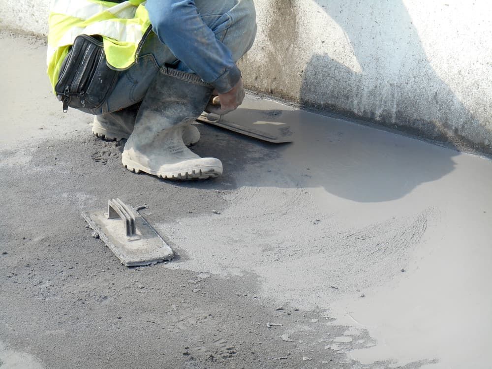 A Man Is Kneeling Down While Using a Trowel on A Concrete Surface — Cemak Constructions in Laurieton, NSW