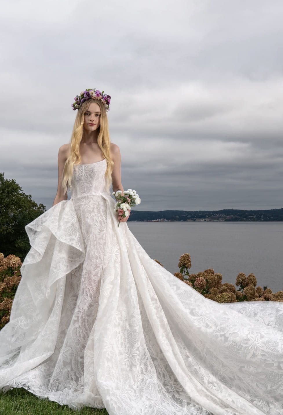 A woman in a wedding dress is standing in front of a body of water holding a bouquet of flowers.