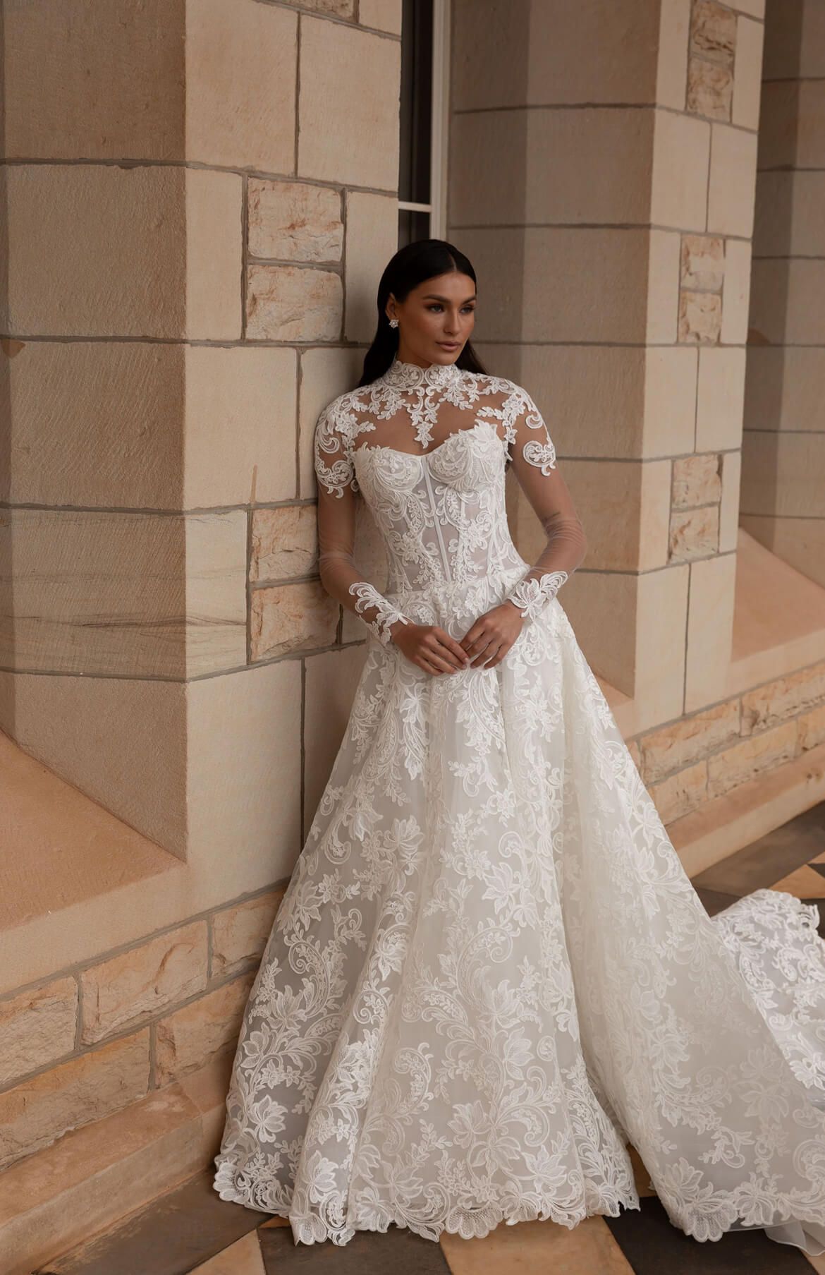 A woman in a white lace wedding dress is leaning against a brick wall.