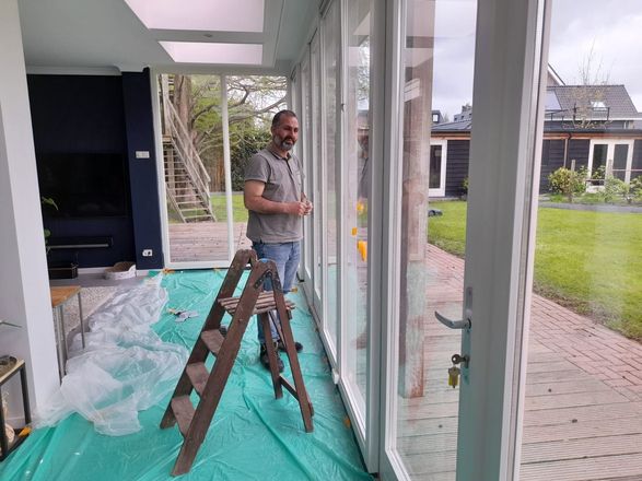 Een persoon staat in een serre met grote glazen deuren en kijkt naar de camera, vlakbij een trapje op een groen afdekzeil.