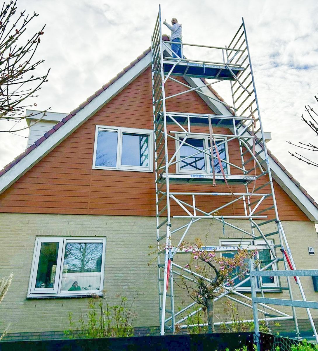 Een persoon die werkt op een steiger voor de kopgevel van een huis met bakstenen en betegelde buitenmuren.