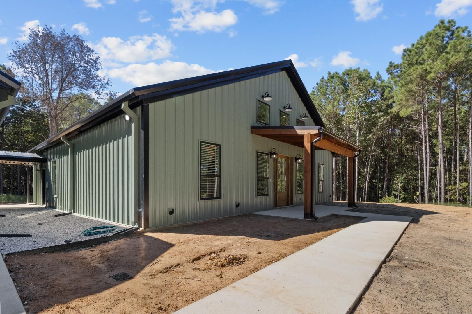 Green metal building with brown porch, concrete walkway, and tall trees against a blue sky.