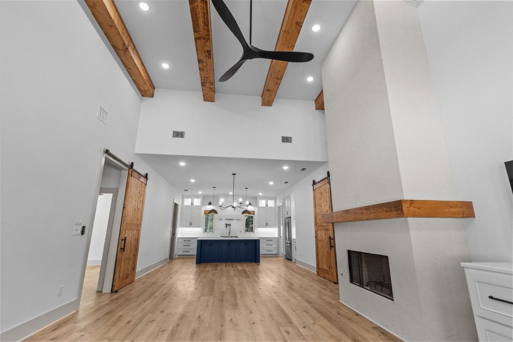 Spacious living room with exposed wooden beams, fireplace, and kitchen in the background.
