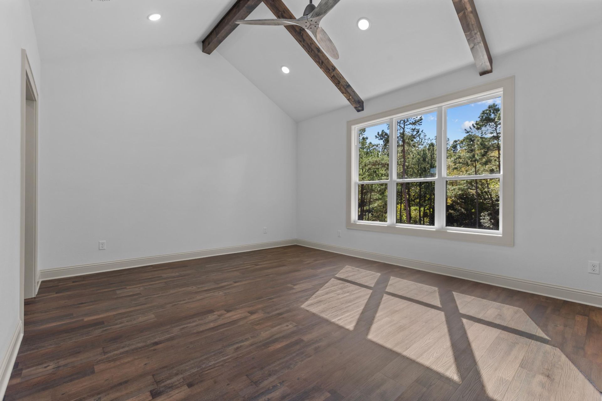 Empty room with hardwood floors, large window, and dark wooden beams.