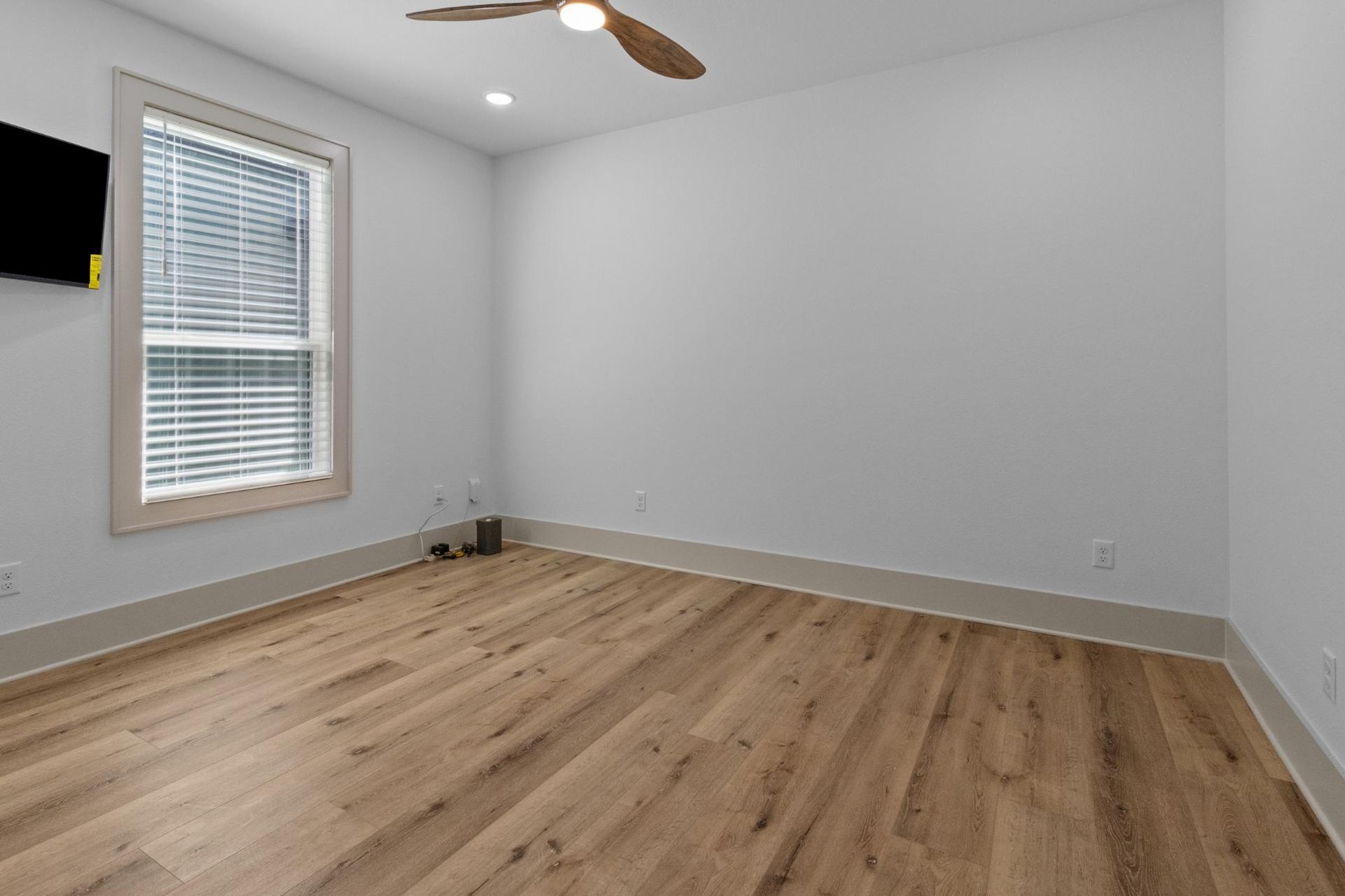 Empty bedroom with light wood floor, white walls, window with blinds, and ceiling fan.