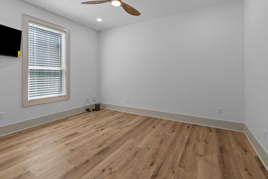 Empty bedroom with light wood floor, white walls, window with blinds, and ceiling fan.