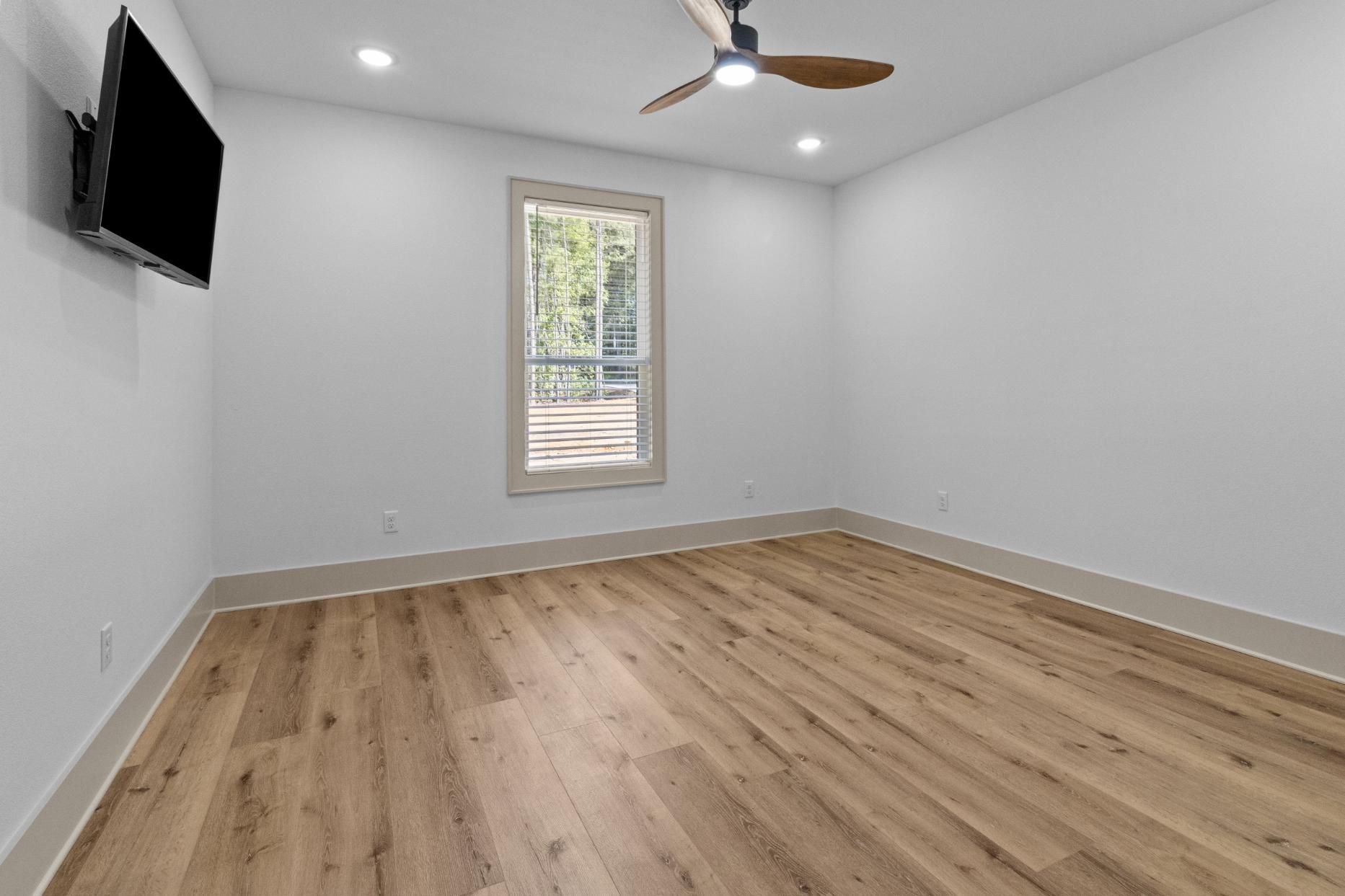 Empty room with wood floors, white walls, a window, ceiling fan, and a mounted TV.